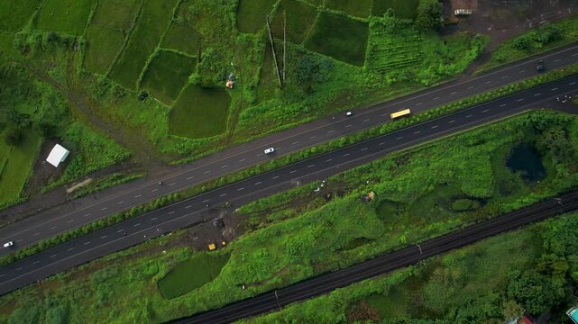 Top-down drone view of a divided highway and parallel railway tracks crossing a lush green rural landscape.