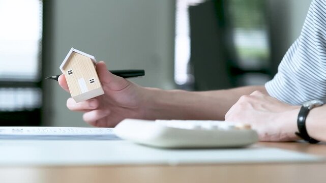 Person holding a wooden house model while calculating costs and reviewing documents, concept of home buying, mortgage, property investment and financial planning.