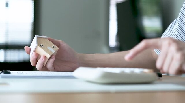 Person holding a house model and using a calculator to plan housing expenses, concept of mortgage calculation, home purchase, budgeting, finance and real estate investment.