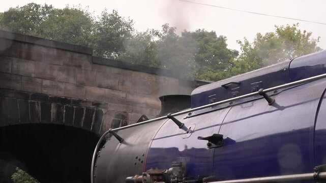Blue steam locomotive boiler moves beneath an old stone bridge as white smoke drifts upward on an overcast day.