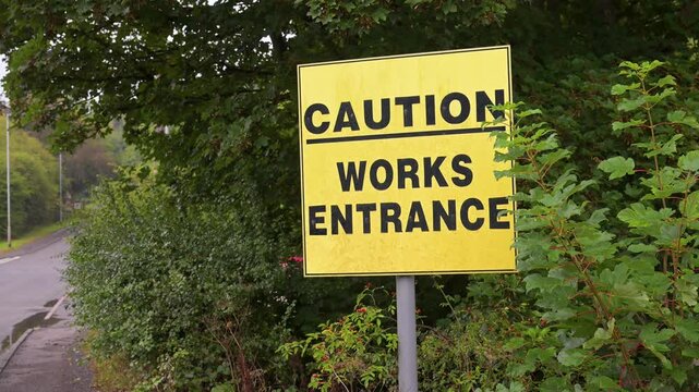 Bold yellow Caution Works Entrance sign stands beside a quiet roadside bordered by dense green foliage.
