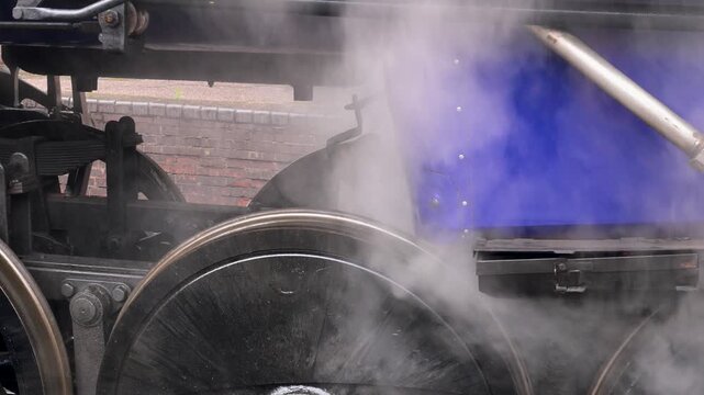 Thick steam swirls around the driving wheels and blue side panel of an LMR steam locomotive in tight close up.
