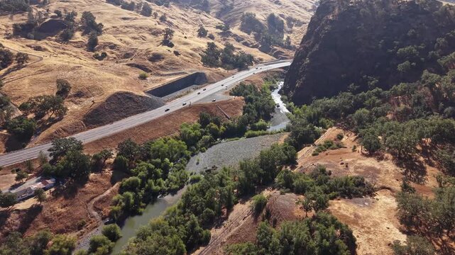 High-angle aerial perspective of traffic driving along a scenic highway next to the Russian River and Frog Woman Rock, set against a backdrop of rolling golden hills in Northern California