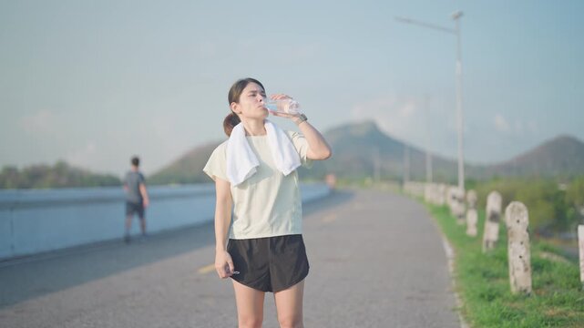 Hot Asian woman with towel drinking water from a reusable bottle after jogging in the park. Exhausted female runner refreshing after summer fitness workout. Hydration and heatstroke concept