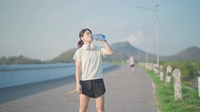Young Asian woman with headphones drinking water from a reusable bottle after jogging in the park. Female runner refreshing after summer fitness workout. Hydration and sustainability concept