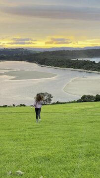 A girl runs on the grassy meadow towards a stunning orange sunset over the bay at Minnamurra Lookout, near Jervis Bay, Wollongong and Kiama, New South Wales, Australia.