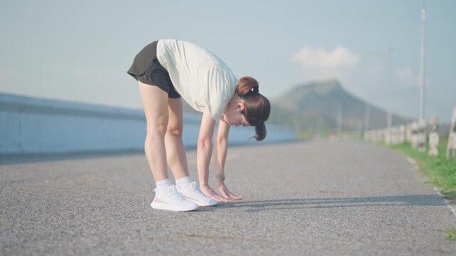 Asian woman with a towel around her neck cooling down and stretching after jogging in the park. Female runner relaxing after morning workout. Fitness, recovery and healthy lifestyle concept