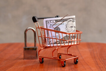 Miniature shopping cart filled with US dollar banknotes next to a steel padlock on a wooden table, secure online shopping and financial data protection concept. © Isman