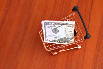Miniature shopping cart filled with US dollar banknotes on a polished wooden table, consumer spending and retail inflation concept with copy space. © Isman