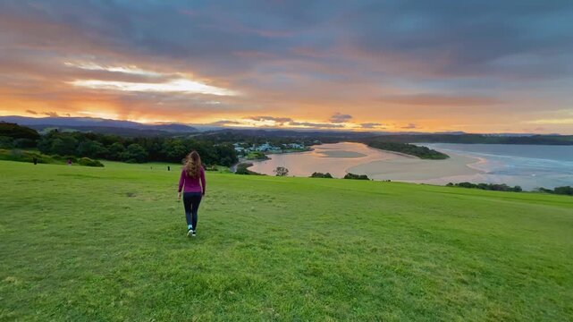 A girl walks on the grassy meadow admiring a stunning orange sunset over the bay at Minnamurra Lookout, near Jervis Bay, Wollongong and Kiama, New South Wales, Australia.