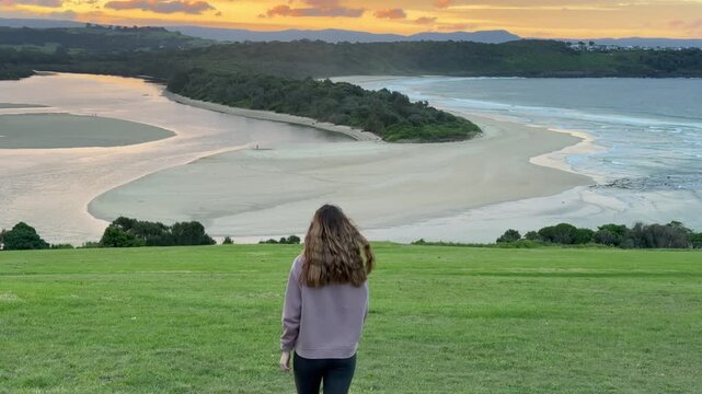 A girl walks on the grassy meadow admiring a stunning orange sunset over the bay at Minnamurra Lookout, near Jervis Bay, Wollongong and Kiama, New South Wales, Australia.