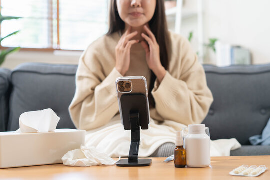Sick Asian woman holding medicine pill blister pack during online video call telehealth consultation with doctor via smartphone on sofa at home