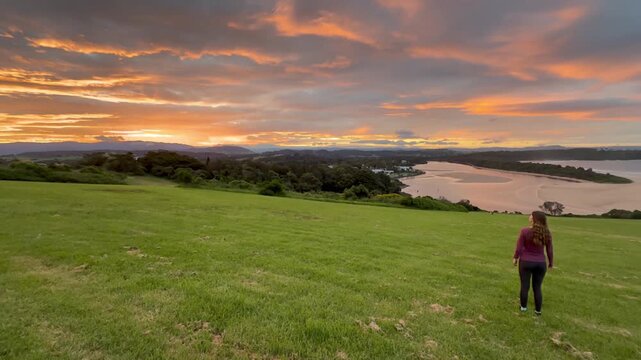 A girl stands on the grassy meadow admiring a stunning orange sunset over the bay at Minnamurra Lookout, near Jervis Bay, Wollongong and Kiama, New South Wales, Australia.