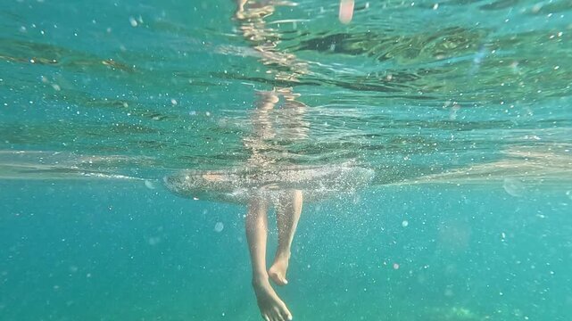 Swimmer dangling legs into crystal blue sea from inflatable ring, underwater perspective. Split shot tracking movement.