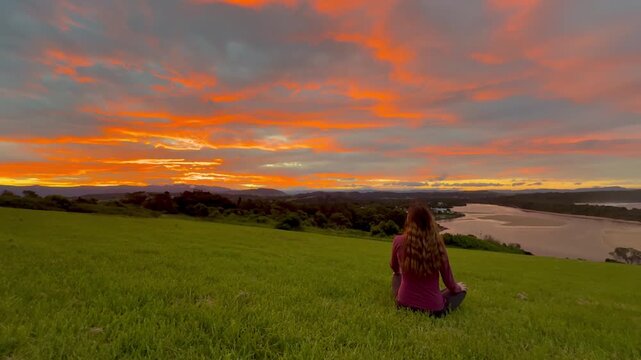 A girl sits on the grassy meadow admiring a stunning orange sunset over the bay at Minnamurra Lookout, near Jervis Bay, Wollongong and Kiama, New South Wales, Australia.
