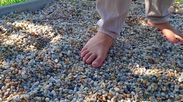 A low angle camera shot shows a child walking barefoot on a gravel sensory path to stimulate nervous system health and physical reflexology in rural Als&oacute;mocsol&aacute;d, Baranya County, Southern Hungary.