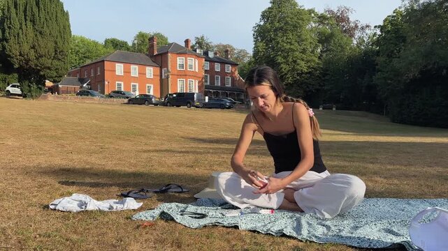 Woman sitting on blanket outdoors, showing joker playing cards on hotel lawn in summer sunlight