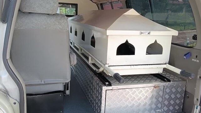 White Muslim funeral coffin or casket inside a funeral transport vehicle, featuring pointed-arch decorative openings inspired by Islamic architecture, a peaked lid, and a metal diamond-plate platform.