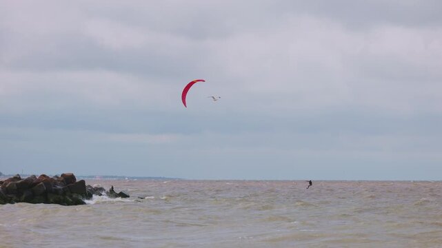 Action wide shot of a lone kitesurfer with a red kite gliding across rough ocean waters on an overcast day. Extreme water sports and active lifestyle concept. Flying over water in slow motion..