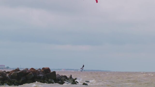 Action wide shot of a lone kitesurfer with a red kite gliding across rough ocean waters on an overcast day. Extreme water sports and active lifestyle concept. Flying over water in slow motion..