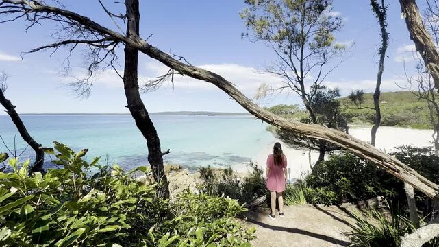 A girl in a pink dress admires the turquoise ocean and white sand at wild Chinamans Beach, on the White Sands Walk near Greenfield Beach, Jervis Bay, New South Wales, Australia.