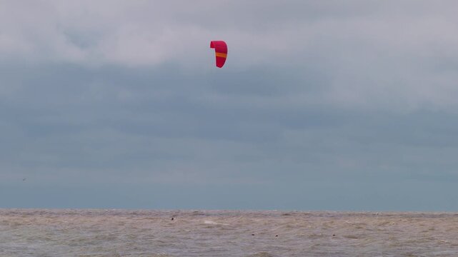 Action wide shot of a lone kitesurfer with a red kite gliding across rough ocean waters on an overcast day. Extreme water sports and active lifestyle concept. Flying over water in slow motion..