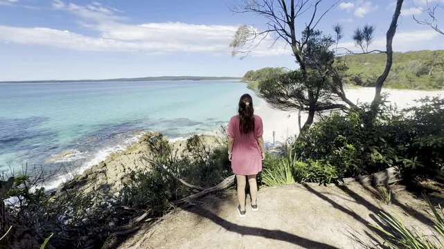 A girl in a pink dress admires the turquoise ocean and white sand at wild Chinamans Beach, on the White Sands Walk near Greenfield Beach, Jervis Bay, New South Wales, Australia.
