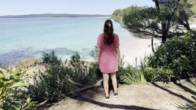 A girl in a pink dress admires the turquoise ocean and white sand at wild Chinamans Beach, on the White Sands Walk near Greenfield Beach, Jervis Bay, New South Wales, Australia.
