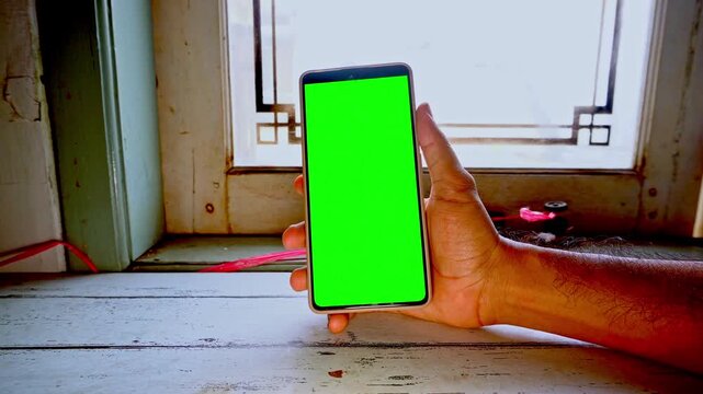A hand holding a mobile phone with a bright green screen for chroma key compositing in a rustic room setting with a wooden window frame backdrop during a clear daylight session for projects.