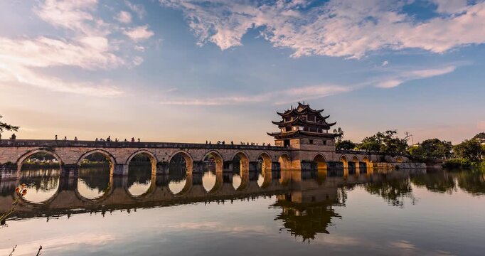 Time-lapse of Shuanglong Bridge at sunset, Jianshui, Yunnan, China. Ancient stone bridge with Chinese pavilion, water reflection and flowing clouds