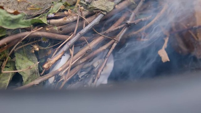 carefully blows on burning paper beneath thin dry twigs to strengthen the flame and start a wood fire in an outdoor fire pit for barbecue preparation. Close up.