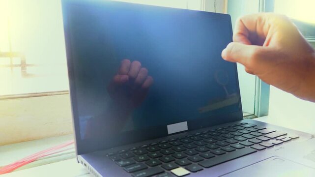 A close up view of a hand reaching out to touch a black laptop screen reflecting the fingers in an office environment to signify digital communication connection and human machine interaction