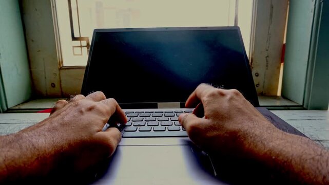 Close up point of view of a person typing on a silver laptop keyboard in a bright office space environment with natural lighting reflecting through the window glass during the productive workday.