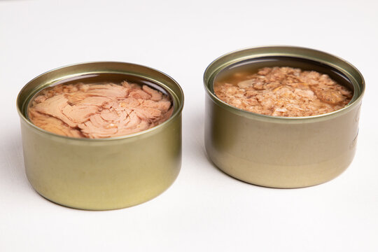 Canned tuna comparison close up. Two open tin cans with solid chunks and shredded flaked fish fillet in diagonal row on white background. Horizontal high angle creative seafood photography with copy s