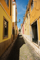 Fototapeta premium Yellow residential facades and a traditional streetlamp line a sunlit cobblestone alley in Lisbon, Portugal.