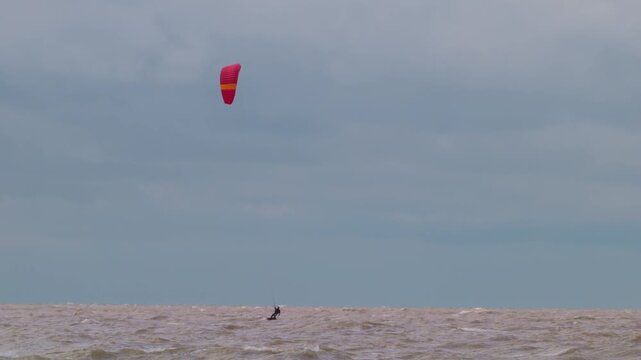 Action wide shot of a lone kitesurfer with a red kite gliding across rough ocean waters on an overcast day. Extreme water sports and active lifestyle concept. Flying over water in slow motion..