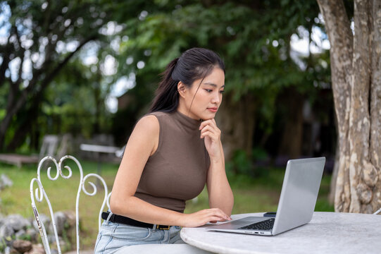 Thoughtful Asian Freelancer Using Laptop At Outdoor Garden Cafe