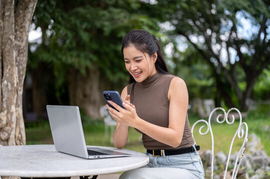 Smiling Asian Woman Texting On Smartphone With Laptop Outdoors