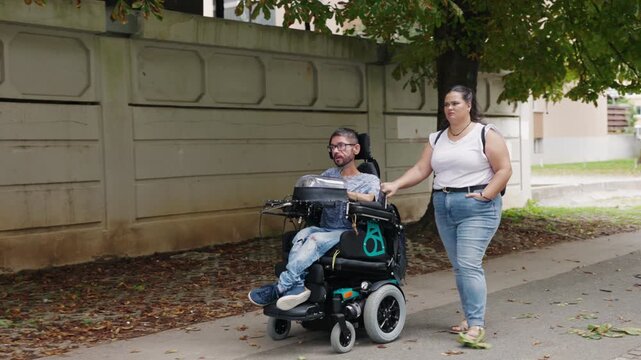 Personal assistant accompanying a powered wheelchair user during an everyday outdoor stroll on an accessible street sidewalk.