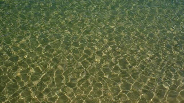 Crystal clear water ripples create intricate dancing patterns of sunlight and shadow on the sandy seabed below. The transparent sea surface reflects the natural daylight, showcasing a serene and vibra