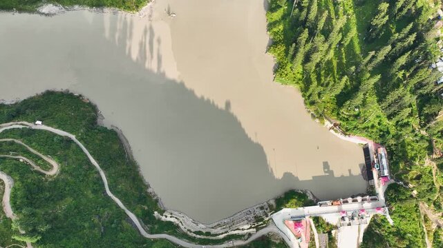 Cinematic drone reveal of the muddy reservoir and dam structure surrounded by dense pine forests, an essential b-roll asset for environmental documentaries on Himalayan rivers.