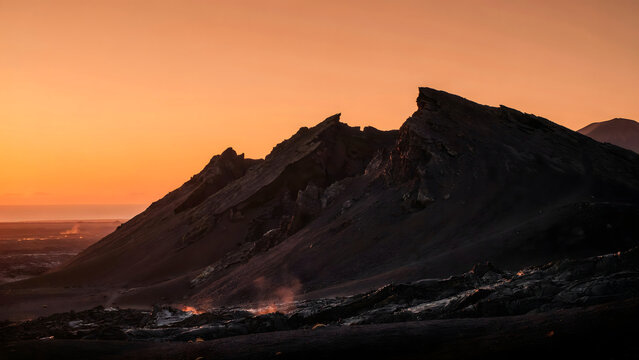 Sharp volcanic mountain ridge silhouette against a bright orange sunset sky with wisps of steam rising from the cooling lava field landscape