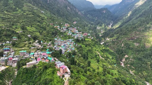 Aerial orbiting shot of traditional mountain guest houses clinging to steep, lush slopes in the high-altitude Himalayan settlement of Tosh, India.