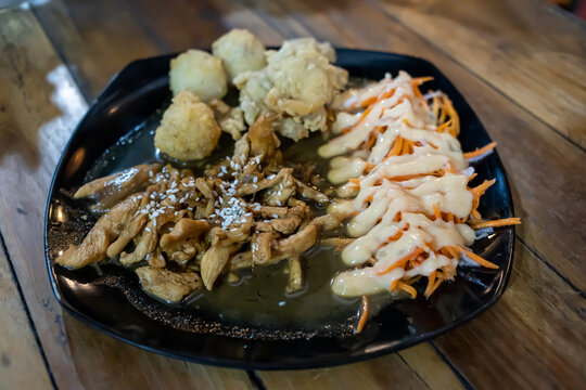 Close up shot of sliced chicken teriyaki topped with sesame seeds, fresh carrot salad with mayonnaise, and deep fried side dishes served on a dark plate over a rustic wooden table.
