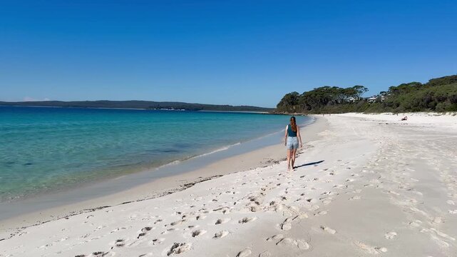 A relaxed girl walks along the white sand of Chinamans Beach, with turquoise water, gentle waves and surrounding bush, near Wollongong, Jervis Bay, New South Wales, Australia.
