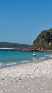 A relaxed girl walks along the white sand of Chinamans Beach, with turquoise water, gentle waves and surrounding bush, near Wollongong, Jervis Bay, New South Wales, Australia.
