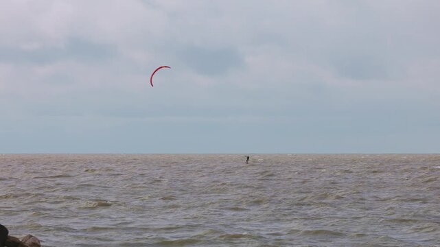Action wide shot of a lone kitesurfer with a red kite gliding across rough ocean waters on an overcast day. Extreme water sports and active lifestyle concept. Flying over water in slow motion..
