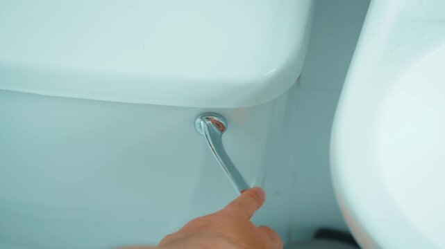 Hand pressing chrome toilet flush lever beside white ceramic tank. Person flushing water after restroom use in bathroom. Toilet hygiene, sanitation routine and household plumbing in close up