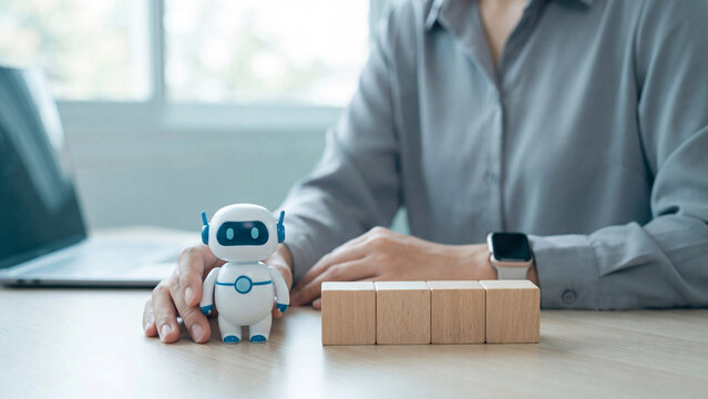 AI integration and workflow automation concept, person arranging a white and blue toy robot beside four blank wooden blocks on an office desk with a laptop and a smartwatch visible on their wrist