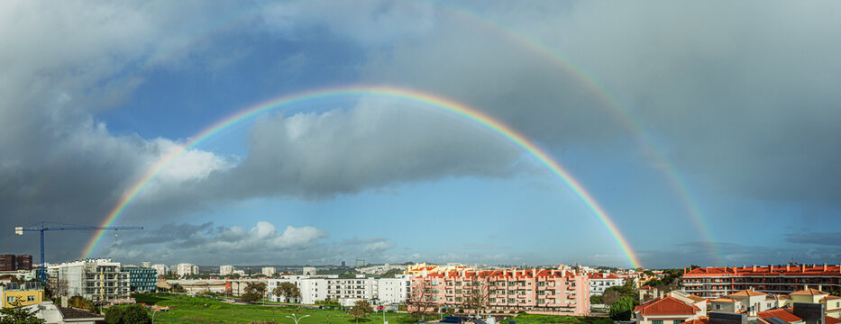 Panoramic view of a double rainbow arching over a residential urban area after rain, with dramatic clouds and sunlight in the sky.